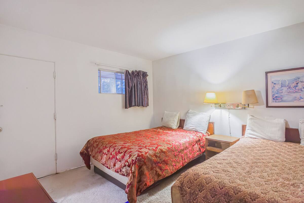 Hotellihuone, jossa on kaksi sänkyä, ikkuna, jossa on verhot, yöpöytä, jossa on lamppu, ja seinällä oleva kehystetty kuva.