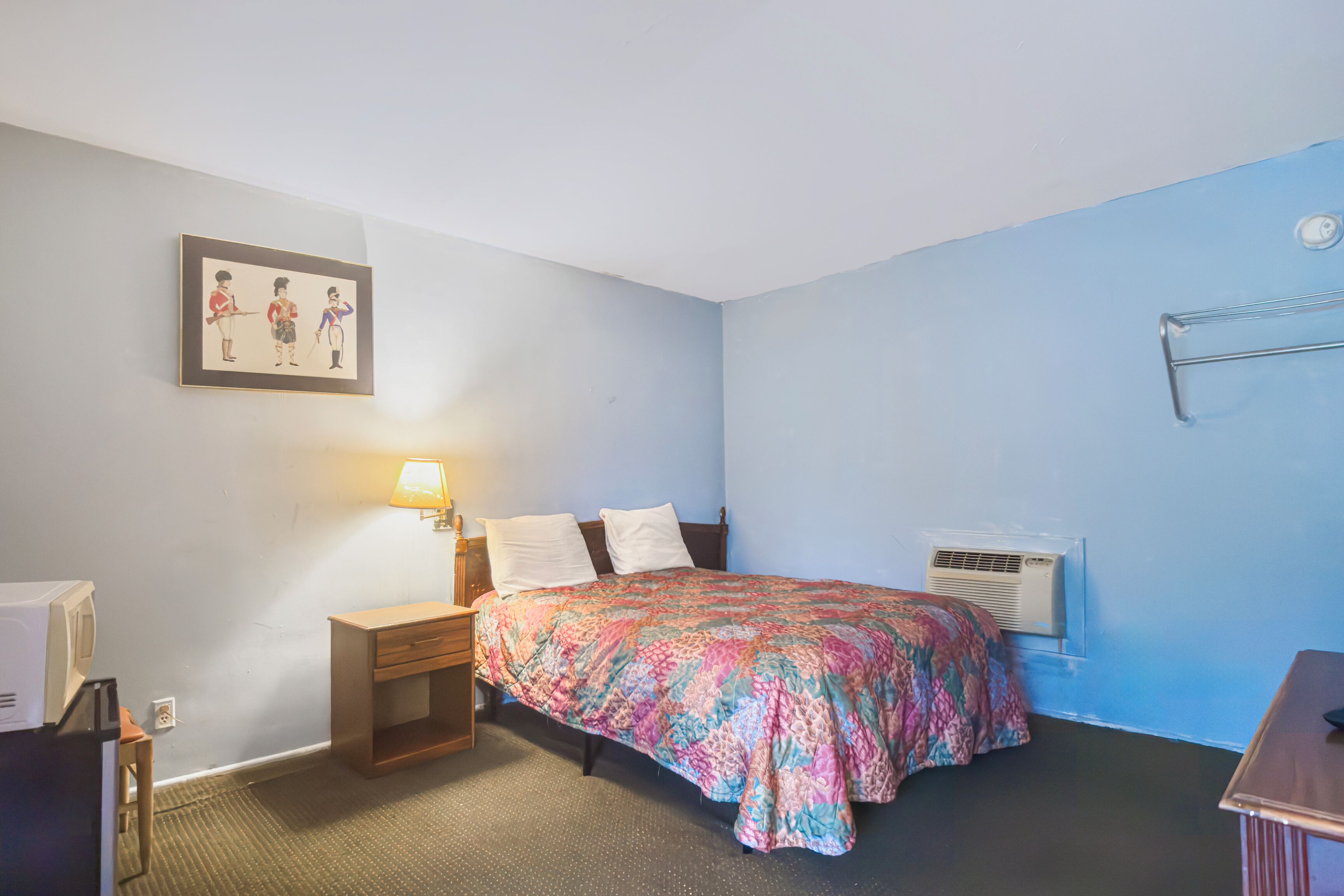 Un dormitorio con cama, mesita de noche, lámpara, aire acondicionado y un cuadro en la pared.