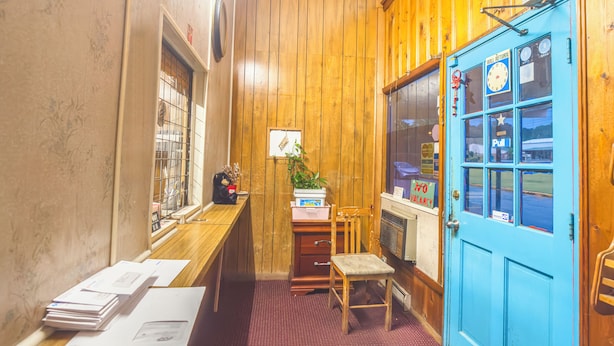 A room with wooden walls, a blue door, a desk with papers, a chair, and a radiator. - Heart of Dixie Motel Near Auburn AL (Dadeville)