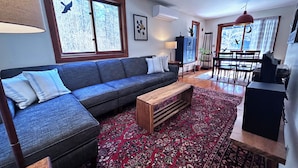 Ett vardagsrum med en grå soffa, ett soffbord i trä, en röd mönstrad matta, en matplats med ett bord och stolar samt ett stort fönster med utsikt över träd.