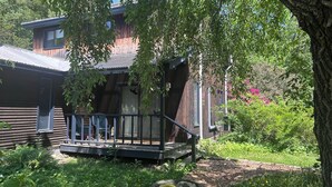 En stuga av trä med en veranda, omgiven av frodig grönska och ett träd.