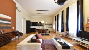A modern living room with a sofa, coffee table, and TV. There is a dining area with chairs and a table. The room has large windows with curtains.
