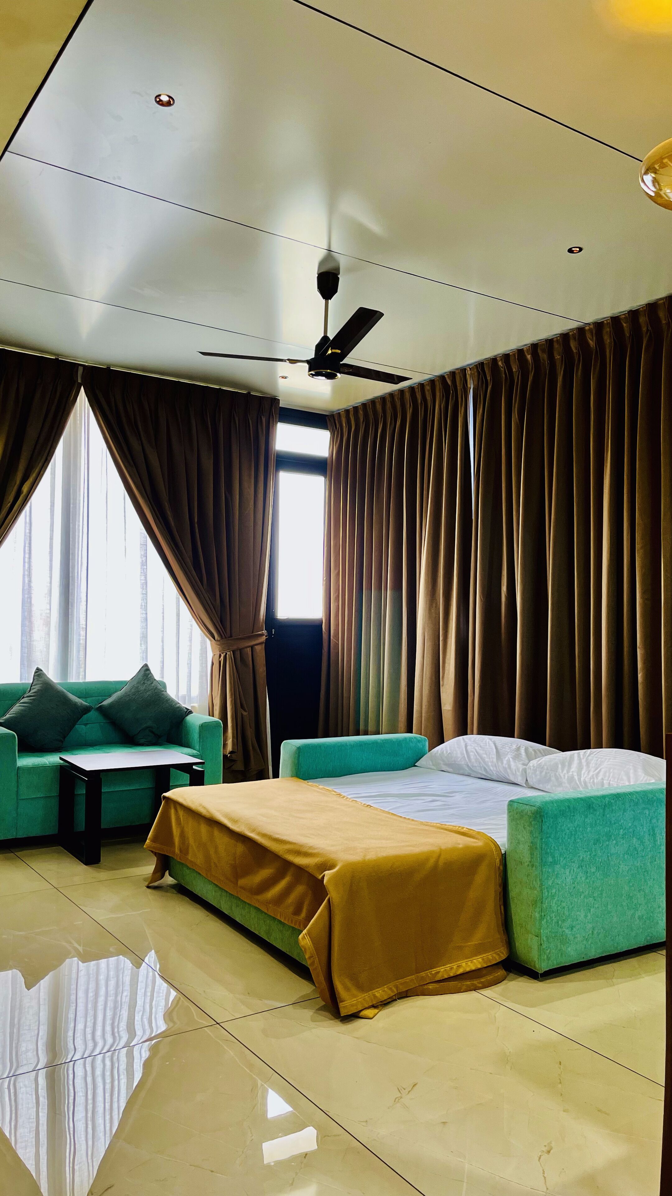 Une chambre d’hôtel moderne avec un lit, deux canapés verts, une petite table et un ventilateur de plafond.
