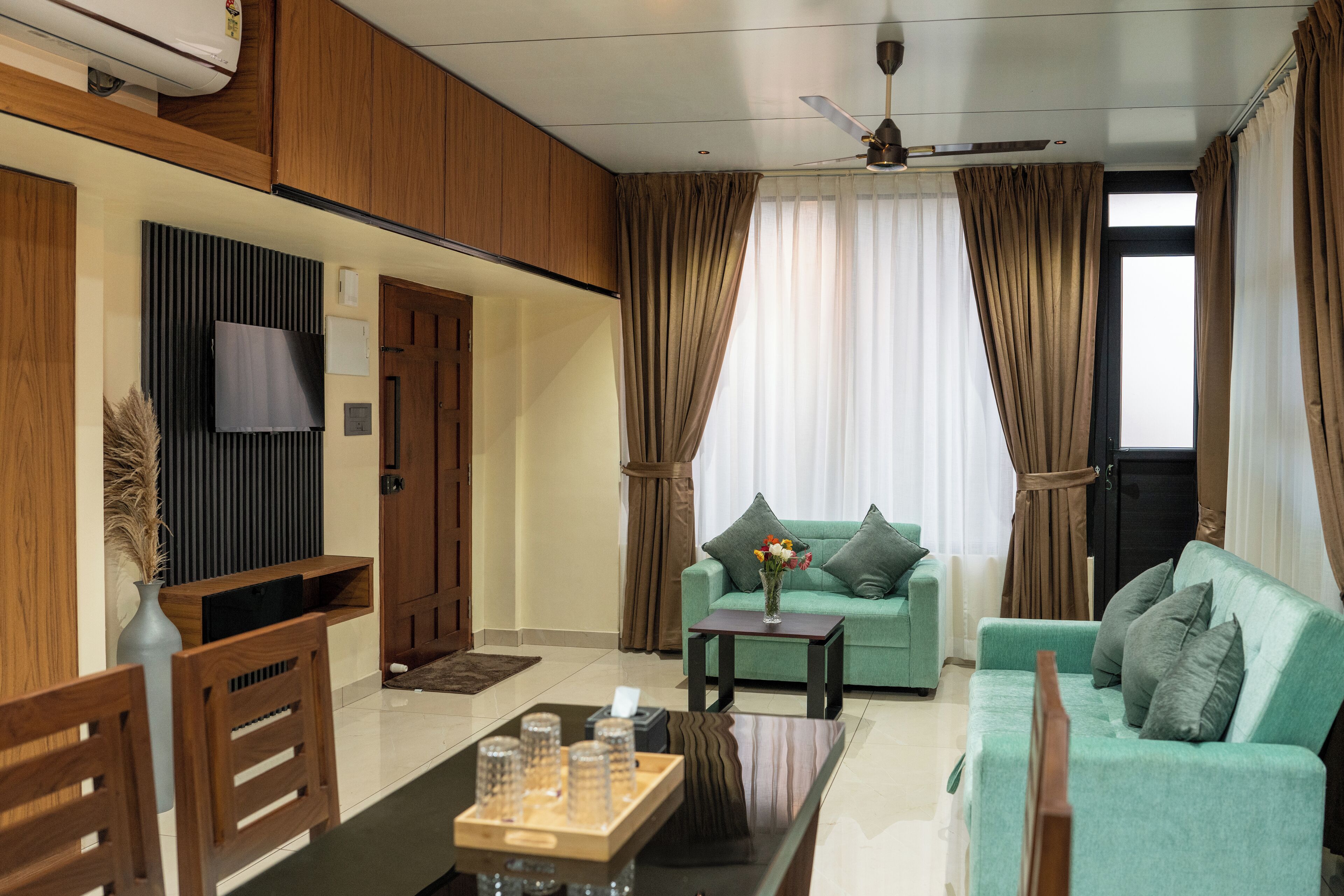 A modern living room with a green sofa, a coffee table, and wooden furniture. There is a TV mounted on the wall and a ceiling fan.