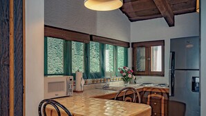 Een rustieke keuken met een houten tafel, barkrukken, een hanglamp, een koelkast en grote ramen met uitzicht op groen.