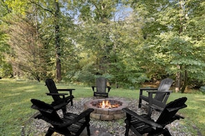 A circular stone fire pit with three Adirondack chairs around it, set in a grassy area with trees in the background. - River View Dog Friendly Game Room w Hot Tub (Sevierville)