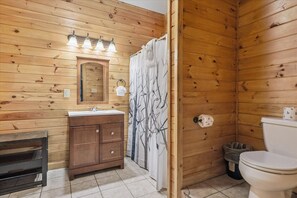 A wooden-paneled bathroom with a wooden bench, a sink with a mirror, a toilet, and a shower curtain with a tree design. - River View Dog Friendly Game Room w Hot Tub (Sevierville)