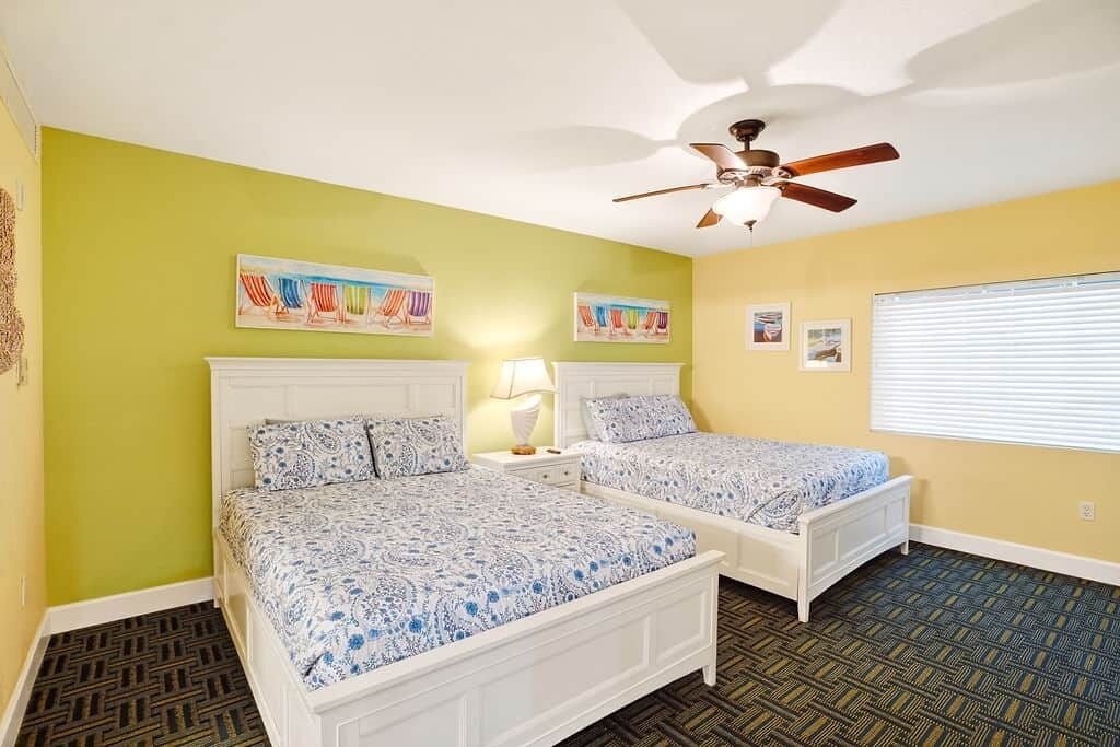 A bedroom with two beds, a ceiling fan, artwork on the walls, and a window with blinds.