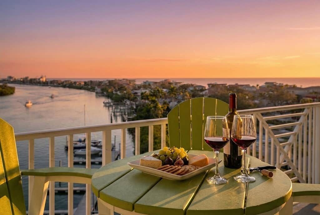 A table with a bottle of wine, two glasses, a plate of cheese and grapes, and a corkscrew on a green chair overlooking a river with boats.