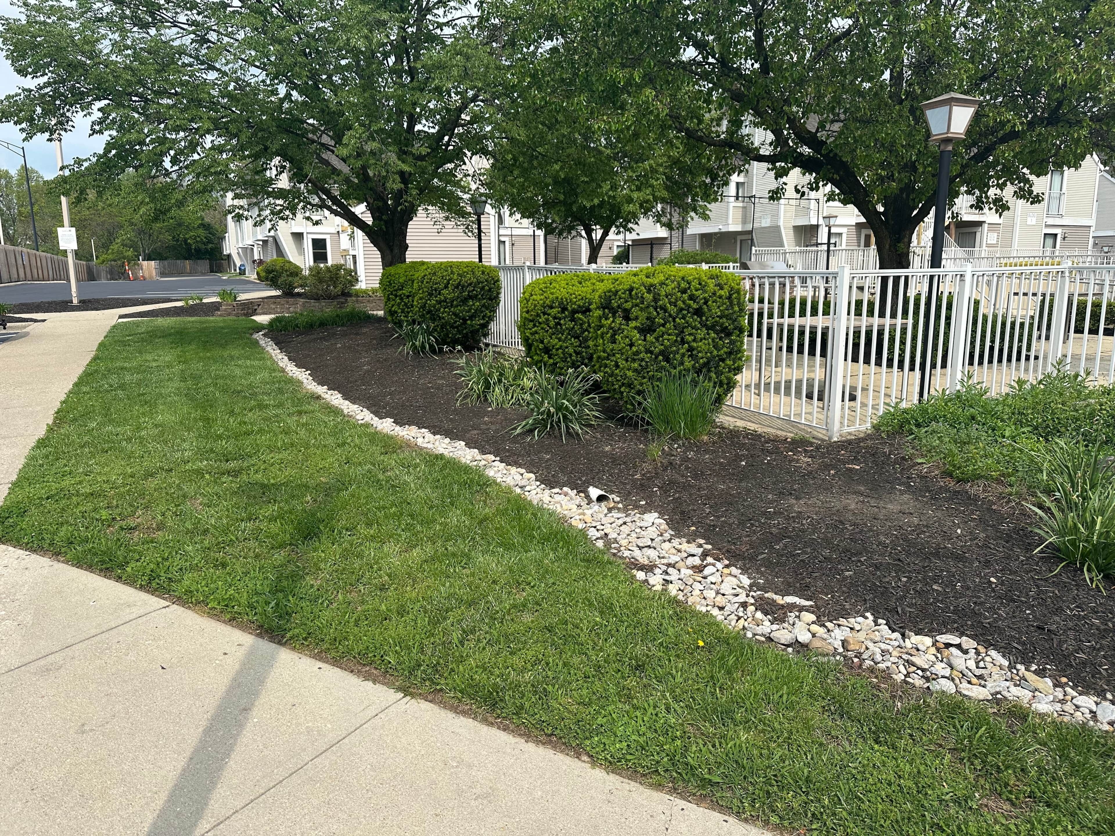 A residential area with a well-maintained lawn, a white fence, and mature trees.