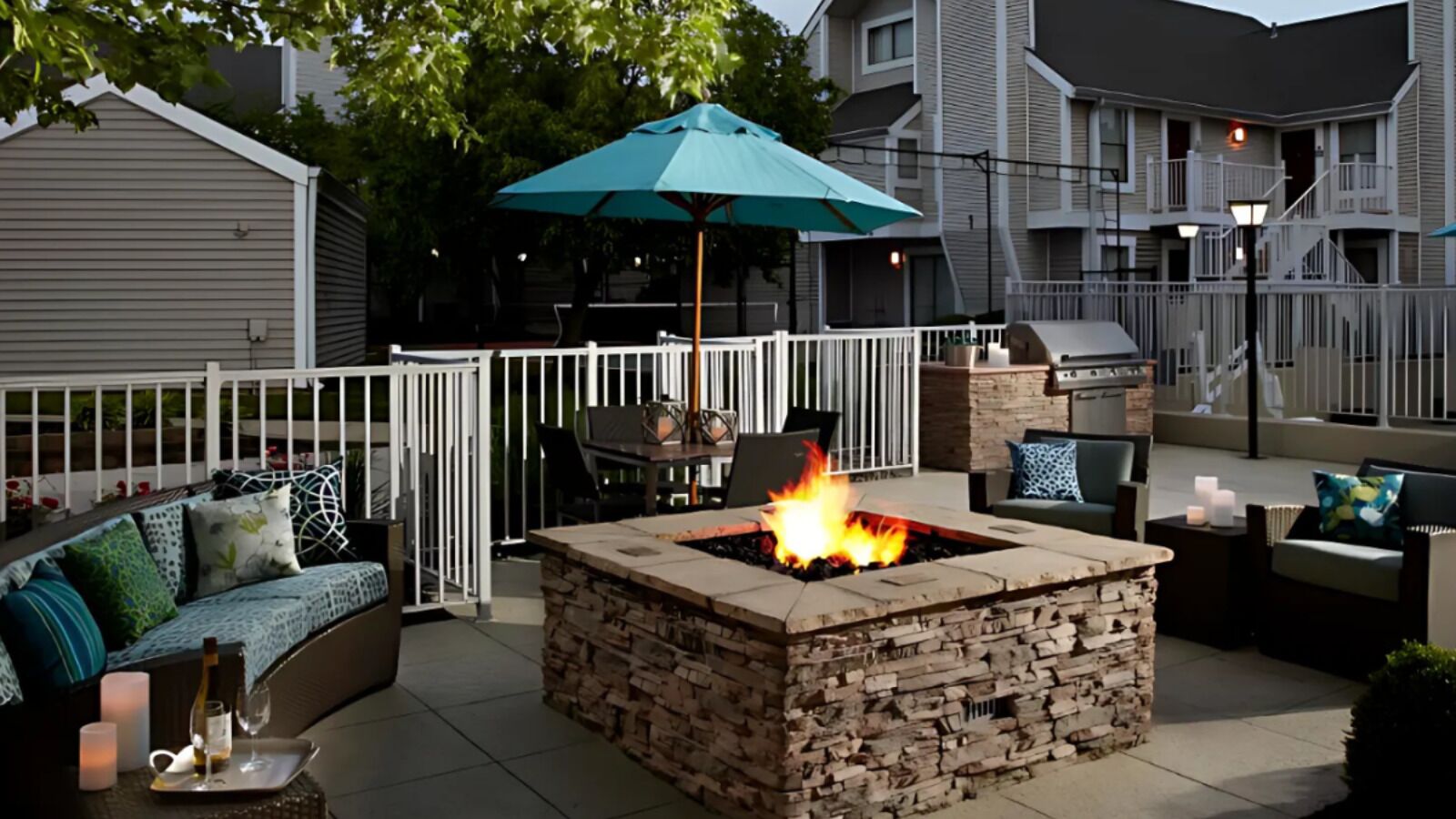 An outdoor patio with a stone fire pit, a white fence, and a blue umbrella.