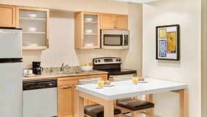 A compact kitchen with a microwave, stove, and dishwasher, a small dining table with two chairs, and a single cabinet with a frosted glass door. - Fully Furnished Studio | MTM | All-Inclusive (Sharonville)