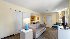 Habitación de hotel con sofá, escritorio con computadora, lámpara y una kitchenette al fondo.
