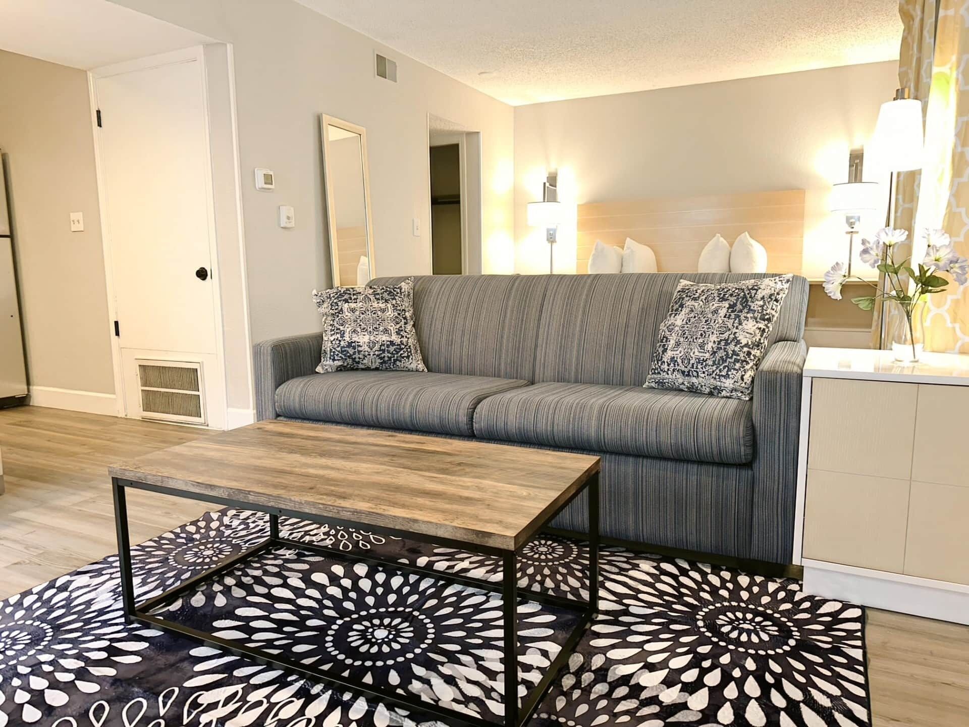 Ein Wohnzimmer mit grauem Sofa, gemusterten Kissen, einem Holztisch und einem schwarz-weißen Teppich.
