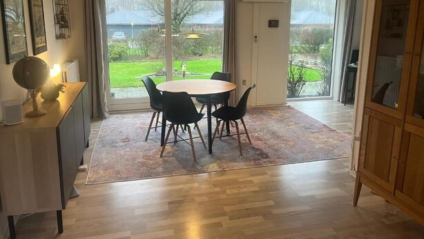 Un comedor con una mesa redonda y sillas, un armario de madera y vistas a un jardín a través de amplios ventanales correderas.
