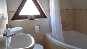 Ein Badezimmer mit Badewanne, Toilette und Waschbecken. Über der Toilette befindet sich ein Fenster mit dreieckigem Rahmen.