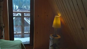 Ein gemütliches Zimmer mit schrägverkleideter Holzbalkendecke, einem großen Fenster mit Blick auf eine verschneite Landschaft, einer mit Grünstoff gepolsterten Bank und einer kleinen Lampe auf einem Holzschemel.