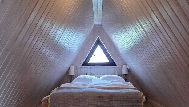Ein gemütliches Schlafzimmer im Dachgeschoss mit schrägen Wänden, einem dreieckigen Oberlicht, einem Holzbett und einem flauschigen Teppich.