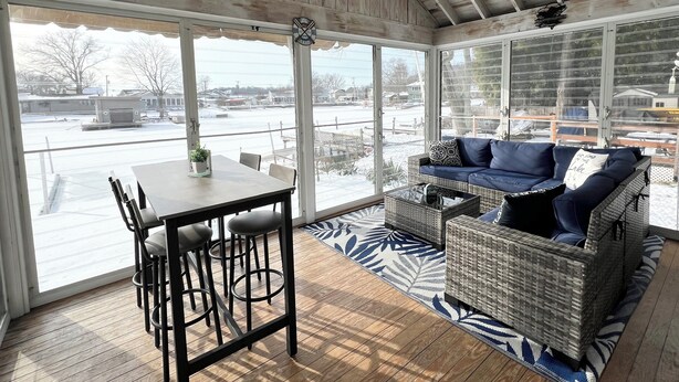 Eine überdachte Terrasse mit Esstisch und Stühlen, ein Korbsessel mit Kissen und Blick auf eine verschneite Landschaft durch große Fenster.