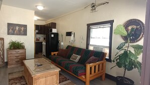 A living room with a sofa, a coffee table, a wooden cabinet, a potted plant, and a kitchen in the background. - Old Hunting Cabin Apartment on High St (Collbran)
