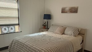 A bedroom with a bed, a nightstand with a lamp, a window with blinds, and a wall-mounted picture. - Old Hunting Cabin Apartment on High St (Collbran)