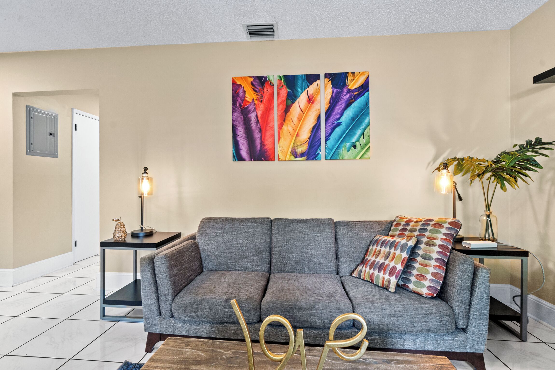 A modern living room with a grey sofa, two side tables with lamps, a colorful feather artwork on the wall, and a 'LOVE' sign on the floor.