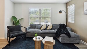 A modern living room with a grey sofa, a black armchair, and a blue rug. There is a plant, a lamp, and a book on a side table.