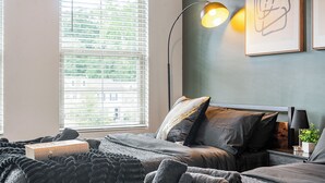 A bedroom with a bed, a bedside table with a lamp, a window with blinds, and a framed artwork on the wall.