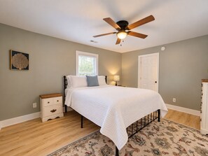 A bedroom with a bed, a nightstand, a ceiling fan, a window, a door, and a patterned rug.