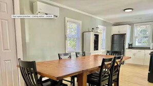 A dining area with a wooden table and black chairs, white cabinets, and a kitchen area with a refrigerator and window blinds.