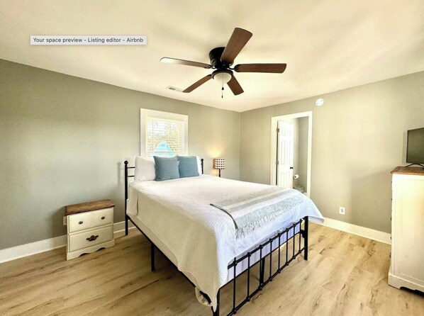 A bedroom with a bed, a nightstand, a bedside table, a ceiling fan, a window with blinds, and a door. - Whispering Shoals - Sleeps 6 - Waterfront home (Beaufort)