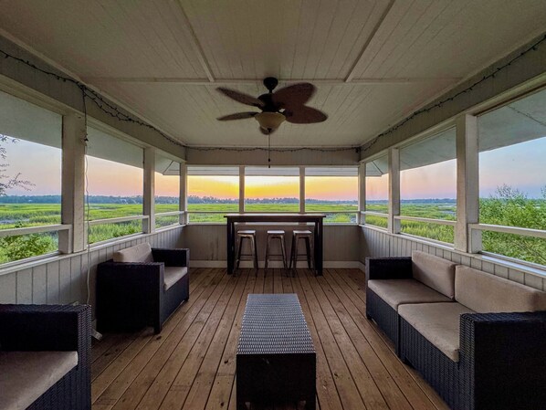A covered porch with wicker furniture, a table, and chairs, overlooking a green landscape. - Waterfront Beaufort Home - Marsh Views & Screened Porch! (Beaufort)