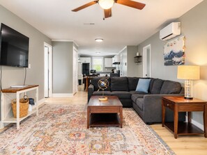 A modern living room with a sofa, coffee table, and TV. There's a kitchen in the background, a patterned rug, and a ceiling fan.