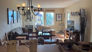 A living room with a sofa, chairs, a coffee table, a chandelier, a television, and a window with a view of a building.