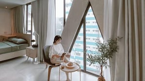 A modern hotel room with a large window, a bed, a reading chair, a small table with a drink, and a potted plant. - Express by M Village Nguyen Van Thu, a brand of Modern Village Lifestyle (Ho Chi Minh City)
