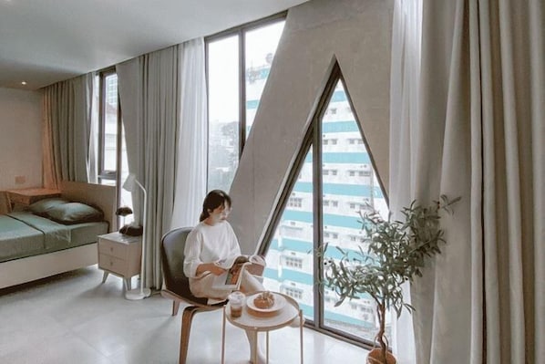Une chambre d’hôtel moderne avec une grande fenêtre, un lit, un fauteuil de lecture, une petite table avec une boisson et une plante en pot.