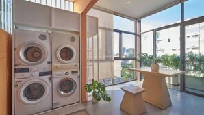 A modern laundry room with four stacked washing machines, a small table with a plant, and large windows offering a view of the city. - Express by M Village Nguyen Van Thu, a brand of Modern Village Lifestyle (Ho Chi Minh City)