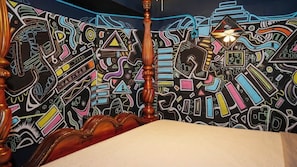 A room with a bed, a wooden headboard, and a wall with colorful abstract patterns.