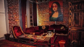 A room with a brick wall, a large painting, a red velvet sofa, a wooden cabinet, and a table with a board game.