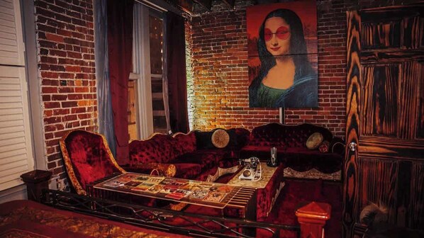 A room with a brick wall, a large painting, a red velvet sofa, a wooden cabinet, and a table with a board game.