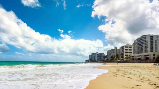 Uma praia com céu claro e ondas do oceano. Prédios ao fundo.