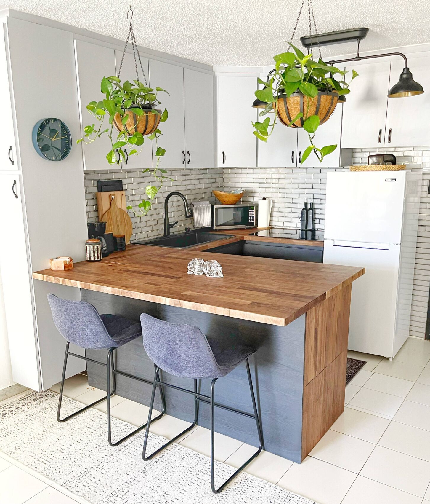 A modern kitchen with a central island, bar stools, hanging plants, and a microwave.