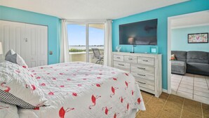 A bedroom with a bed, a dresser, a TV, and a view of the outdoors. - Cozy 2-bedroom condo with heated pool, fitness room, game room, Gulf Front (Hudson)