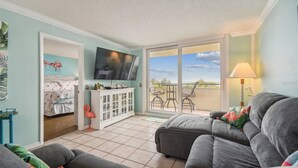 A living room with a grey sofa, a TV on the wall, a balcony with chairs, and a white cabinet. - Cozy 2-bedroom condo with heated pool, fitness room, game room, Gulf Front (Hudson)