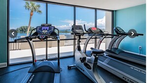 A gym with treadmills and an elliptical machine, large windows with a view of palm trees and water. - Cozy 2-bedroom condo with heated pool, fitness room, game room, Gulf Front (Hudson)