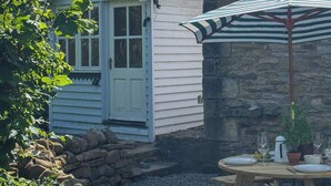 A wooden picnic table with a striped umbrella, a stone wall, and a white building with a blue door.
