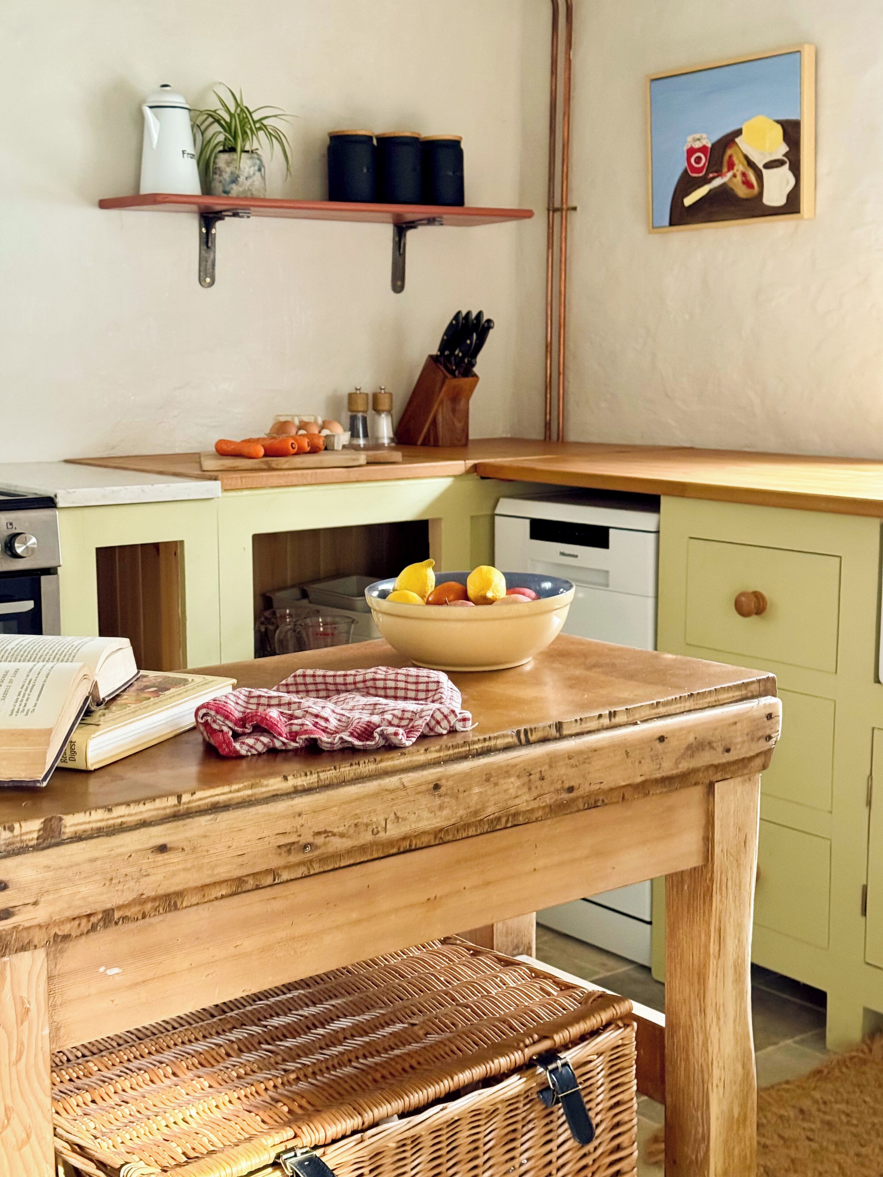 Een keuken met een houten tafel, een fruitschaal, een waterkoker en een schilderij aan de muur.