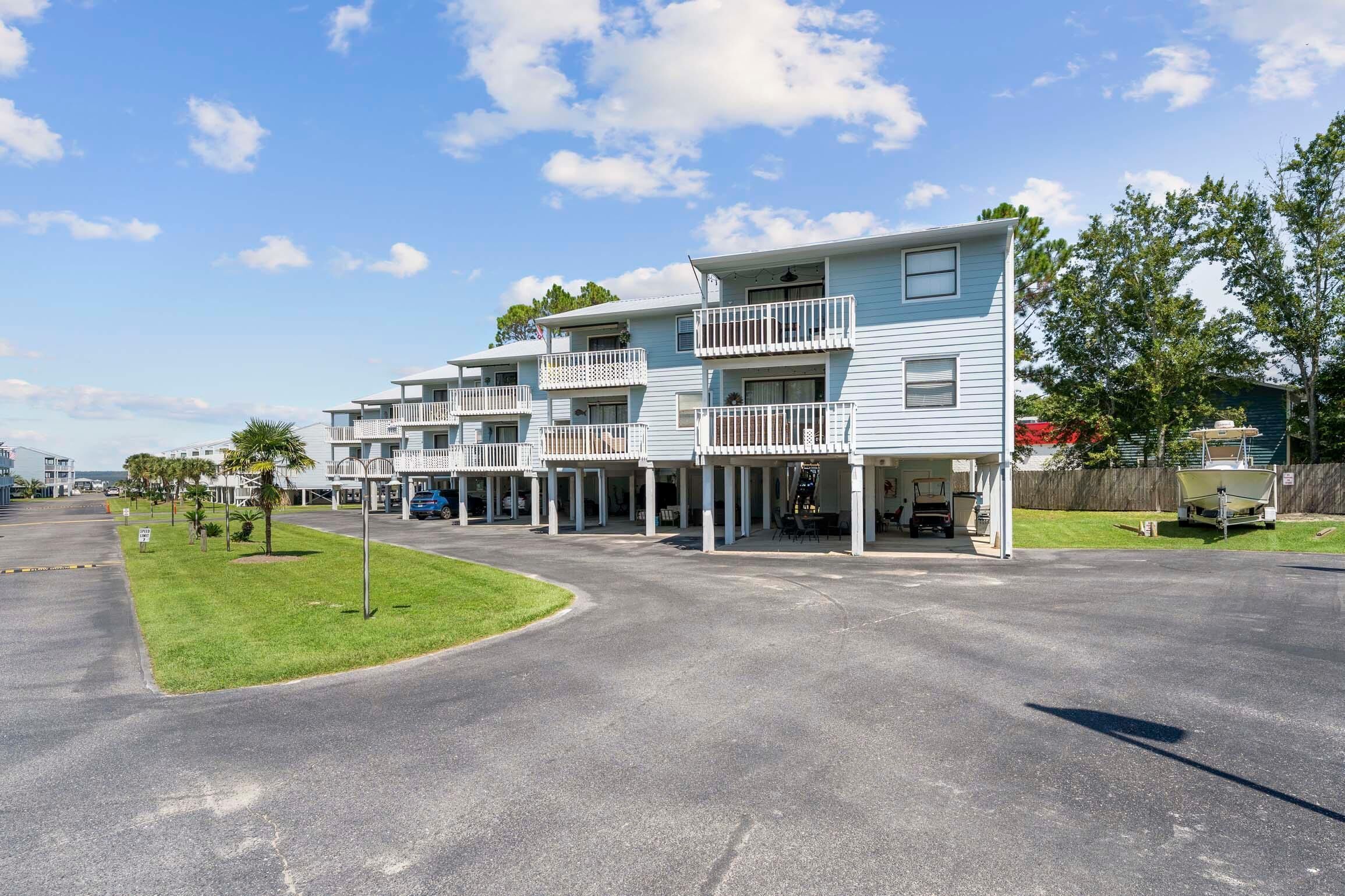 A row of light blue multi-story buildings with balconies, a paved parking area, and a grassy lawn.