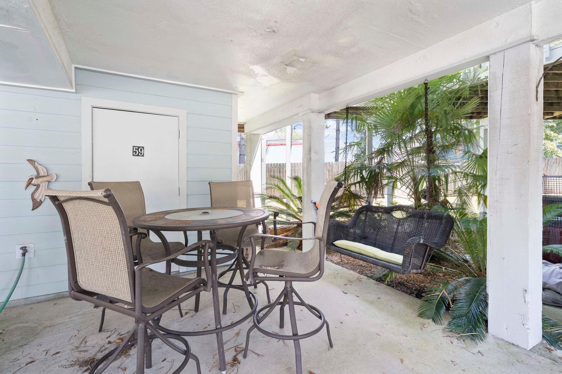 A covered patio area with a table and chairs, a door with the number 59, and a view of palm trees and a fence.