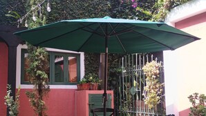 Un patio con una mesa y sillas bajo un paraguas verde, rodeado de plantas en macetas y un edificio rojo.
